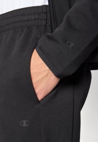 Felpa atletica nera con chiusura a zip, polsini a coste e tasche laterali. Include un dettaglio logo nero sulla manica e nell’area delle tasche.