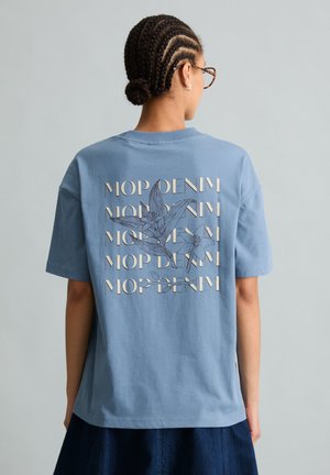 Osoba z warkoczami i okularami, ubrana w niebieską koszulkę z powtarzającym się tekstem "MOP DENIM" i kwiatowymi ilustracjami na plecach.