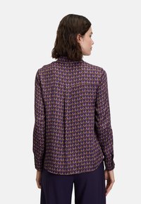 Purple blouse featuring a geometric pattern in orange and blue, with long sleeves and a buttoned collar, made from a smooth fabric.