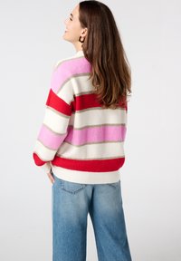 Multicolor gestreepte trui met rode, roze en beige strepen. Gebreid materiaal, wijde mouwen en ribgebreide manchetten en onderrand. Draag met lichtblauwe jeans.