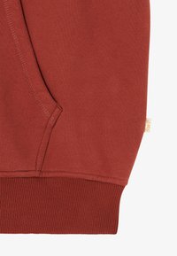 Nærbilde av en rustfarget hettegensers lomme med synlige sømmer og ribbestrikket kant, som har et lite beige stoffmerke på siden.