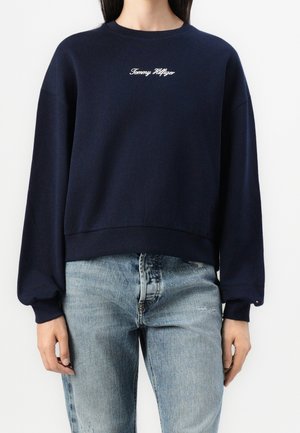 Osoba ubrana w granatową bluzę Tommy Hilfiger i jasnoniebieskie dżinsy, stojąca z rękami luźno opuszczonymi wzdłuż ciała.