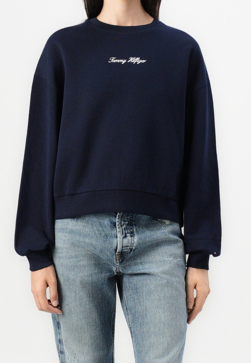 Personne portant un sweat-shirt Tommy Hilfiger bleu marine et un jean bleu clair, debout les mains détendues le long du corps.