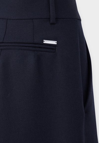Navy trousers with a textured fabric, featuring a discreet silver brand tag and flat front pockets. The waistband is smooth with belt loops.