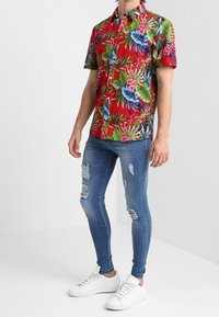 Kurzärmliges rotes Hemd mit Blumenmuster, blauen Blättern und pinken Akzenten; kombiniert mit ausgewaschenen hellblauen Jeans und weißen Sneakers.