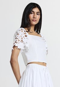 Femme aux longs cheveux noirs portant un haut blanc à manches en dentelle florale et une jupe blanche froncée, devant un fond uni.