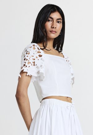 Donna con lunghi capelli scuri che indossa un top bianco con maniche in pizzo floreale e una gonna bianca arricciata su uno sfondo semplice.