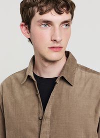 Camicia in velluto a coste marrone con colletto abbottonato, caratterizzata da un tessuto strutturato e una T-shirt nera sotto. Design semplice senza motivi visibili.