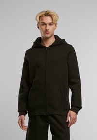 Sudadera con cremallera negra hecha de un tejido suave, que cuenta con una gran capucha, dos bolsillos frontales y puños acanalados. Diseño simple y limpio sin patrones.