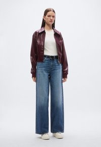 Bordeauxkleurig leren bomberjack, wit T-shirt met ronde hals, wijde blauwe jeans en witte sneakers. Model staat tegen een neutrale achtergrond.