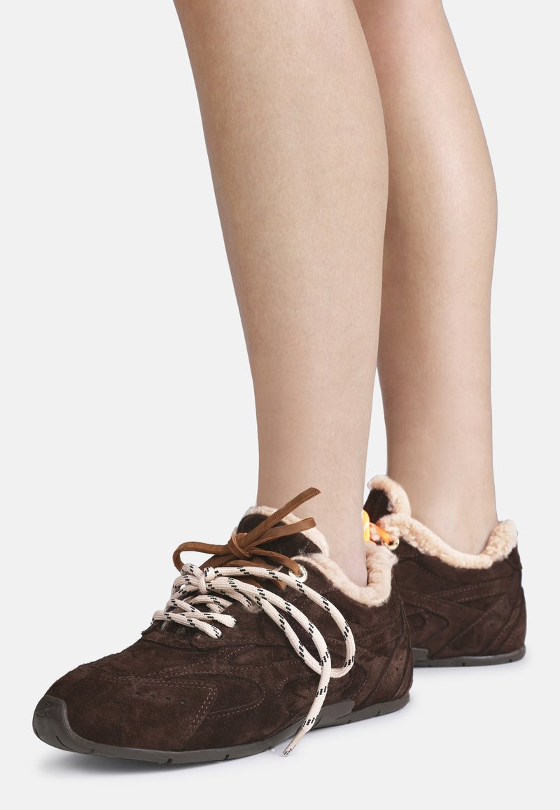 Brune semskede sneakers med lys pelsforing og mismatched snørebånd, båret på nakne ben mot en ensfarget bakgrunn.