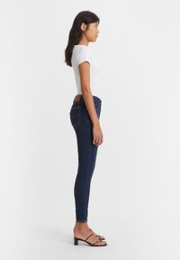 Schmale Jeans aus dunklem Denim mit hohem Bund und Gesäßtaschen. Kombiniert mit einem taillierten weißen T-Shirt und schwarzen, gerafften Absatzschuhen.