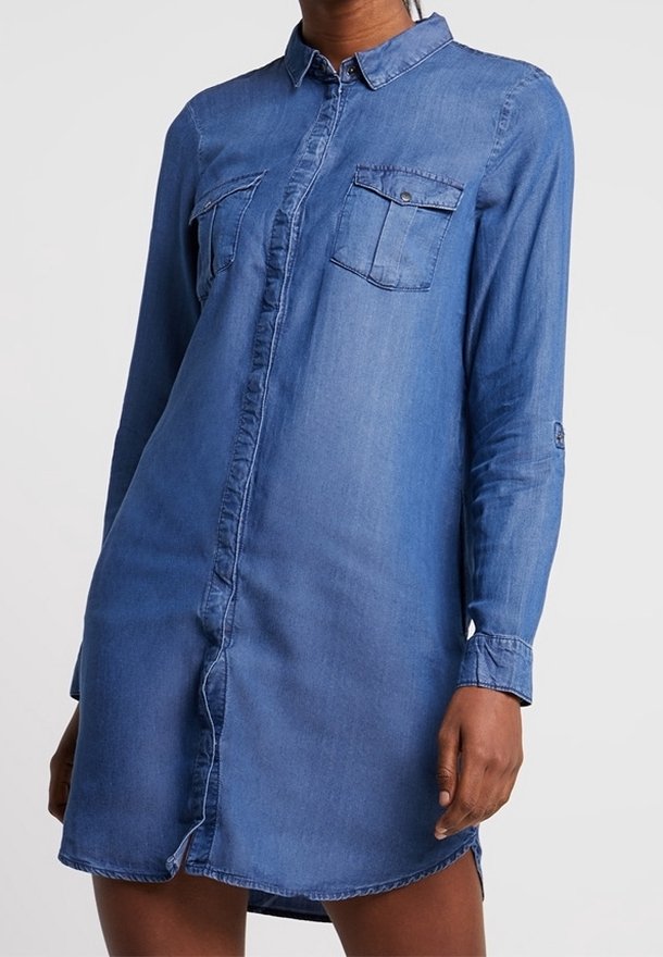 Femme portant une robe chemise en denim bleu à manches longues, boutonnée à l'avant, avec deux poches poitrine et un col.