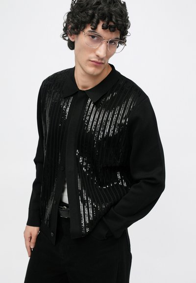 Cárdigan negro con lentejuelas horizontales, diseño con cuello y mangas largas; llevado sobre una camisa blanca y combinado con pantalones negros.