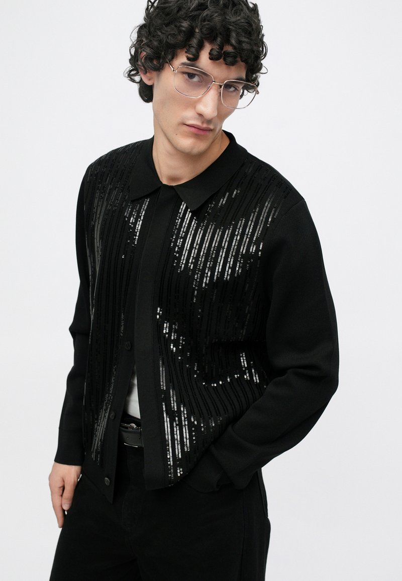 Schwarze Strickjacke mit horizontalen Pailletten, mit Kragen und langen Ärmeln; über einem weißen Hemd getragen und mit schwarzen Hosen kombiniert.