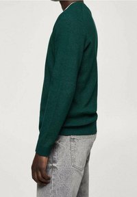 Personne portant un pull à manches longues vert foncé et un jean gris clair, debout de profil devant un fond uni clair.