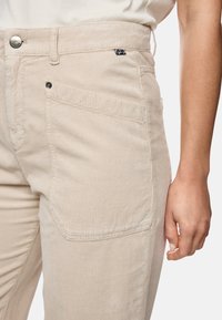 Beige Cordhose aus Kord mit breiten Rippen, ausgestattet mit Vordertaschen, einem Metallknopfverschluss und einem dezenten Markenlogo-Akzent nahe dem Bund.