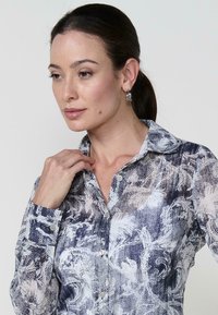 Una donna con una camicetta a fiori tocca il colletto, guardando pensierosa di lato su uno sfondo chiaro.
