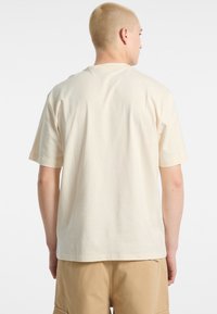 T-shirt di cotone crema con maniche corte, vestibilità rilassata, scollatura tonda e texture liscia, vista dalla schiena, abbinata a pantaloni corti beige.