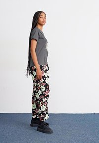 Tricou gri cu design floral, pantaloni negri cu model floral și adidași negri. Modelul stă în profil, pe un fundal simplu.