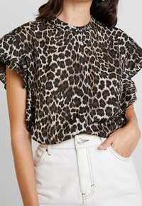 Blouse à manches courtes imprimé léopard en noir, marron et beige, avec un col montant et un tissu froncé autour des épaules. Associée à un jean blanc.