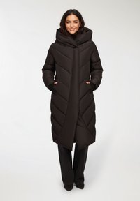 Czarna kurtka puchowa z dużym kapturem, wzorem w zygzak, bocznymi kieszeniami i długim krojem, zestawiona z ciemnymi spodniami i butami na obcasie.