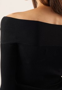 Haut noir côtelé à épaules dénudées avec manches longues, présentant un motif texturé et des transitions de tissu lisses au niveau des coutures des épaules.