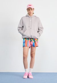 Ljusgrå hoodie med framficka, färgglada mönstrade shorts med blommor och randigt mönster, rosa sneakers och en rosa keps.