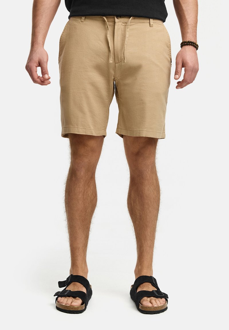 Helle beige Shorts mit einer lockeren Passform, vorderen Taschen und einem Kordelzugbund, kombiniert mit schwarzen Sandalen mit zwei Riemen und gepolsterten Sohlen.