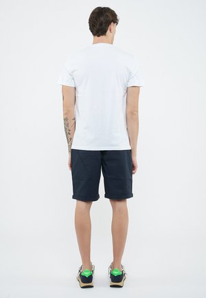 Uomo in piedi di spalle, indossa una maglietta bianca, pantaloncini blu navy con risvolto, tatuaggi sul braccio sinistro e scarpe da ginnastica con tacchi verdi.