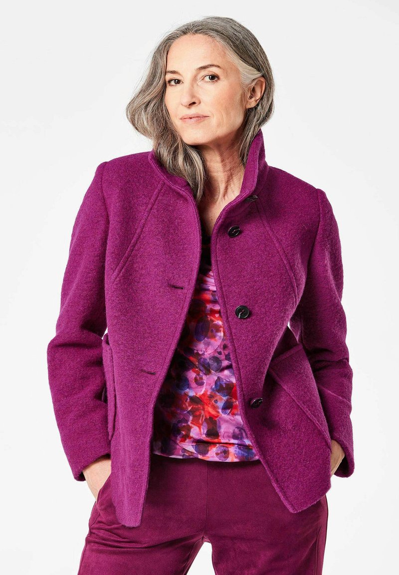 Fuchsia wollen jas met een hoge kraag, drie zwarte knopen en zijzakken, gedragen over een meerkleurige bloemenprint top en bijpassende broek.