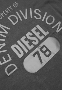 T-shirt grigia con grande testo bianco che recita "PROPRIETÀ DELLA DIVISIONE DENIM" all'interno di un rettangolo arrotondato, con il numero "78" sottostante.