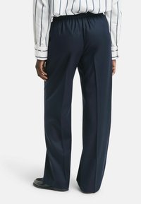 Persoon in marineblauwe wijde broek en een witte shirt met zwarte verticale strepen, staande tegen een effen achtergrond.