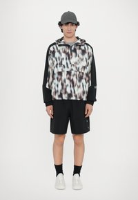Geacă de vânt cu un model abstract alb-negru, buzunar frontal, asortată cu pantaloni scurți negri și adidași albi. Căciulă gri.
