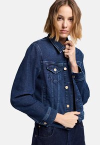 Ciemnoniebieska kurtka dżinsowa z klasycznym kołnierzem, przednimi kieszeniami i złotymi guzikami. Charakteryzuje się usztywnionym fasonem i lekko zwężanymi rękawami.