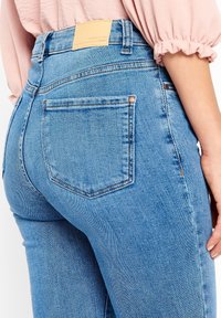 Närbild av en person som bär ljusblå jeans med en bakficka och en beige läderlogotyp, tillsammans med en rosa långärmad topp.