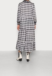 Černobílé kostkované šaty s nabíranými rukávy, stupňovitým designem a volánkovým lemem, doplněné krémovými kozačkami po kolena na neutrálním pozadí.
