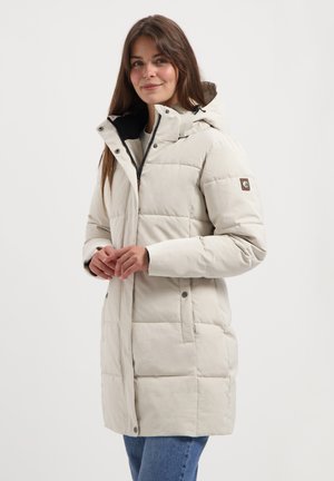 Vrouw in een lichtbeige gewatteerde winterjas met capuchon, zijzakken en een spijkerbroek, staand tegen een effen witte achtergrond.