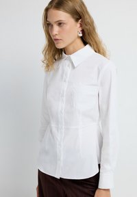 Donna con capelli biondi ondulati che indossa una camicia bianca aderente con taschino e pantaloni a righe scure, in piedi su uno sfondo neutro.