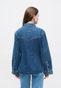 Τζιν μπουφάν σε μέσο μπλε με εφαρμοστή γραμμή. Διαθέτει μυτερό γιακά, μακριά μανίκια και κουμπωτά μανικετόκουμπα. Ραφές με λεπτομέρειες στην πλάτη.