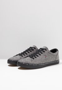 Sneakers in tessuto grigio con lacci neri e suole in gomma. Presentano un logo sul lato e una banda con il marchio lungo il fondo della suola.