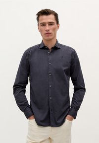 Cămașă button-up de culoare bleumarin închis, cu mâneci lungi, guler ascuțit și un logo subtil pe piept; textură netedă cu croială clasică.