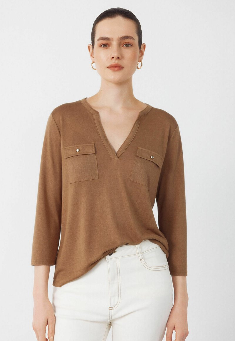 Vrouw draagt een bruin V-hals topje met twee borstzakken en een witte broek, staande tegen een effen lichte achtergrond.