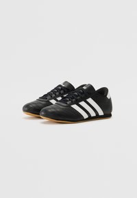 Baskets de sport en cuir noir avec un design à trois bandes blanches, bout rond et surface texturée. Elles possèdent une semelle en caoutchouc et un système de laçage.