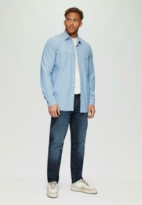 Lichtblauw overhemd met lange mouwen en twee borstzakken, gecombineerd met donkere spijkerbroek en witte sneakers. Eenvoudig, casual ontwerp met knoopsluiting.