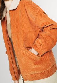 Veste en velours côtelé orange avec un col doux de couleur crème. Elle est équipée d'une fermeture éclair et de poches latérales. Tissu texturé à rayures verticales.
