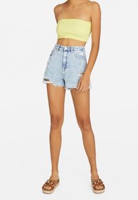 Top sin mangas de color amarillo con fruncidos, pantalones cortos de mezclilla azul claro de tiro alto con detalles desgastados, y sandalias de plataforma marrones con acentos decorativos.