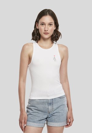 Jeune femme aux cheveux bruns portant un haut blanc sans manches avec un petit motif cœur et main et un short en jean bleu clair.