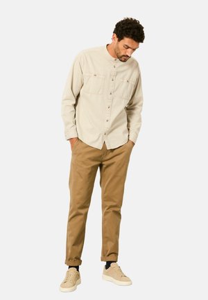 Beiges langärmeliges Hemd mit Stehkragen, zwei Brusttaschen und Knöpfen vorne, kombiniert mit khakifarbenen Chinos und hellen Sneakers.