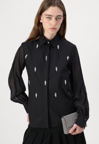 Maglietta nera con maniche a palloncino trasparenti, caratterizzata da decorazioni argentate e chiusura con bottoni. Accompagnata da una clutch argento.